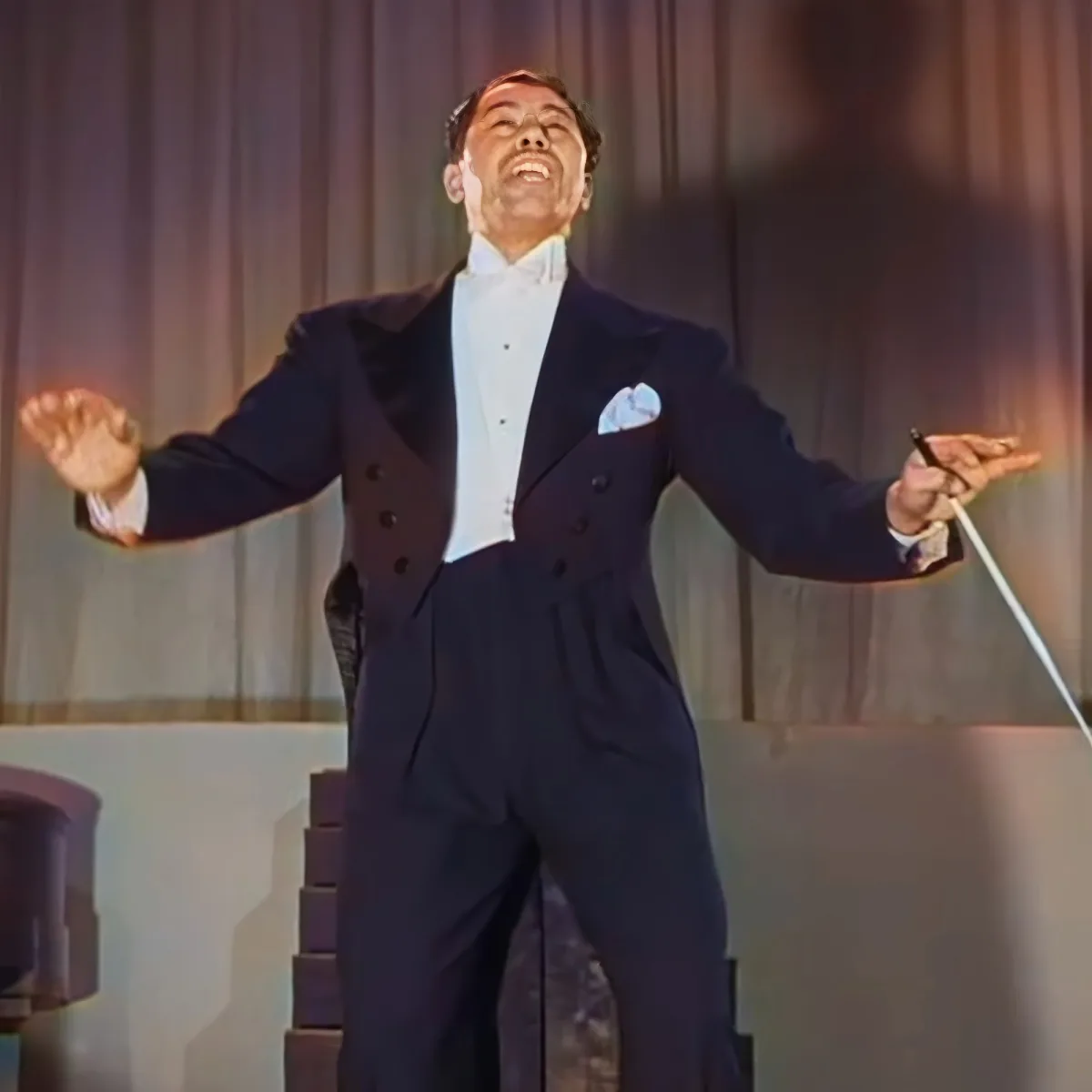 A man in a tuxedo is joyfully singing on stage, holding a cane, with his arms outstretched.