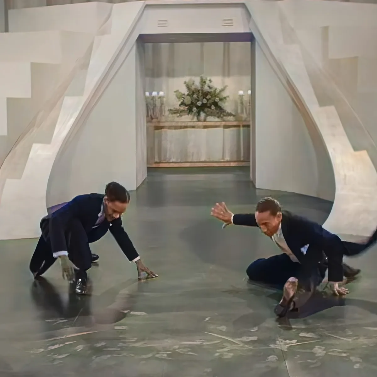 Two men in suits are crouching on a polished floor, seemingly in a race or competition.