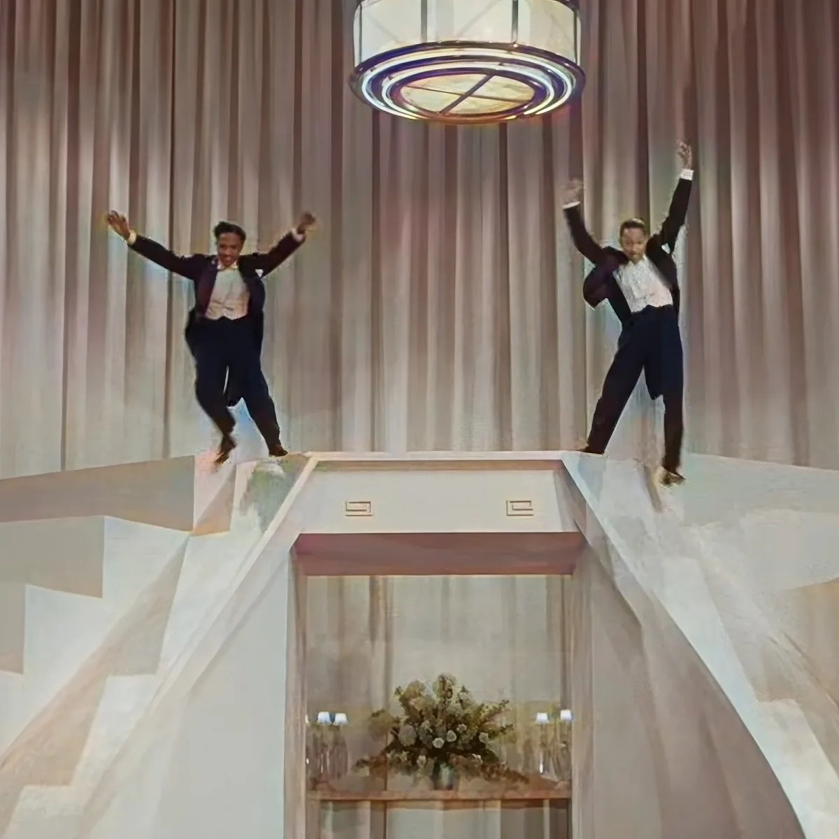 Two men in suits jump joyfully on a raised platform in a grand room with a chandelier and floral arrangement.