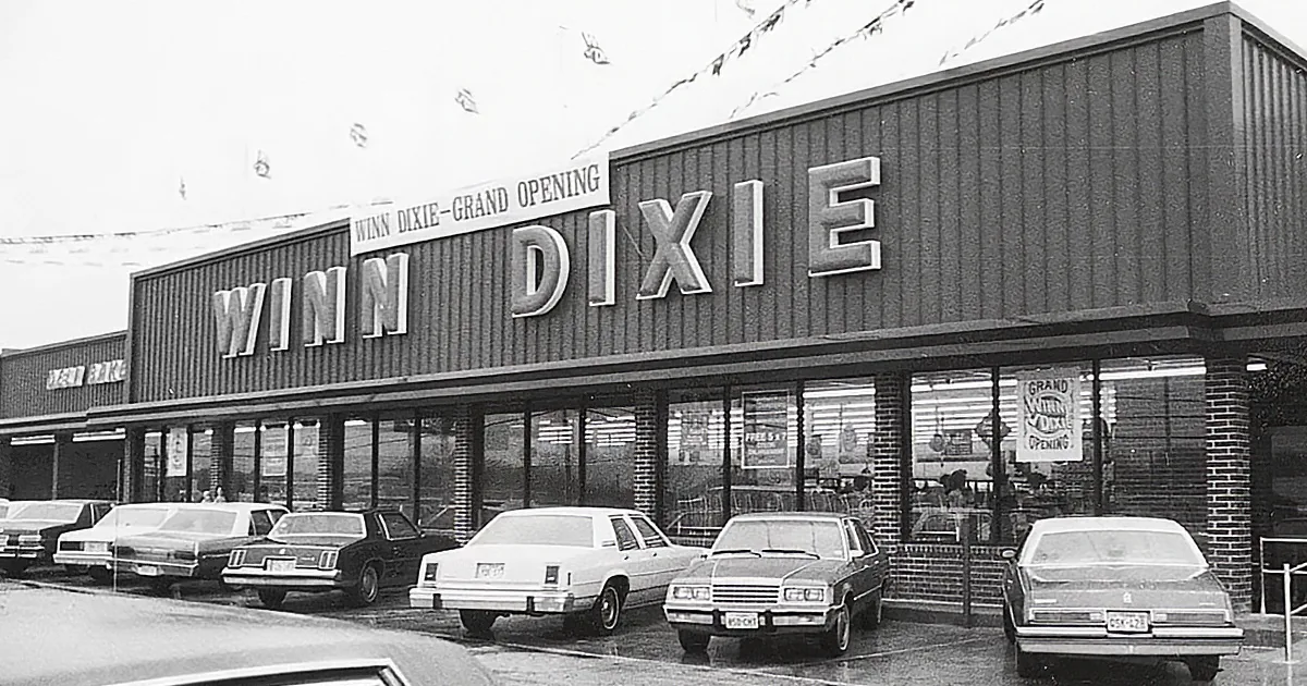 Winn Dixie store with "Grand Opening" banner, cars parked in front, brick facade, and festive decorations.