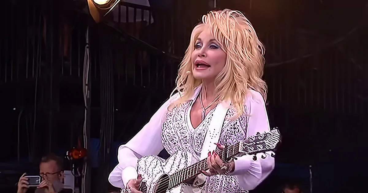 Dolly Parton sings and plays guitar on stage, wearing a white outfit with intricate details.
