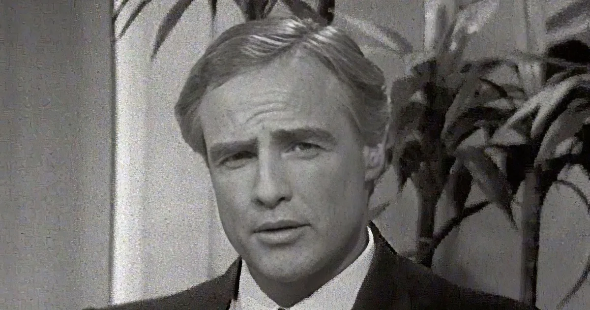 A man in a suit holds a card, smiling, while another man in a turtleneck speaks into a microphone.