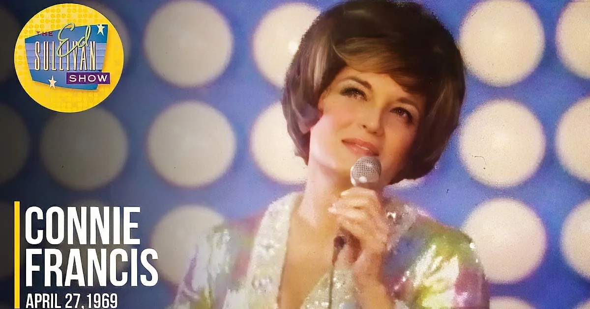 A woman with short hair sings into a microphone, wearing a sparkly top, against a blue background with white circles.