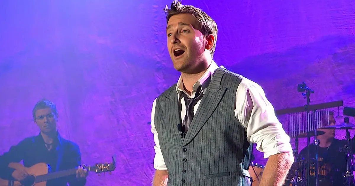 A man in a vest sings passionately on stage, with a guitarist and drummer in the background under purple lighting.