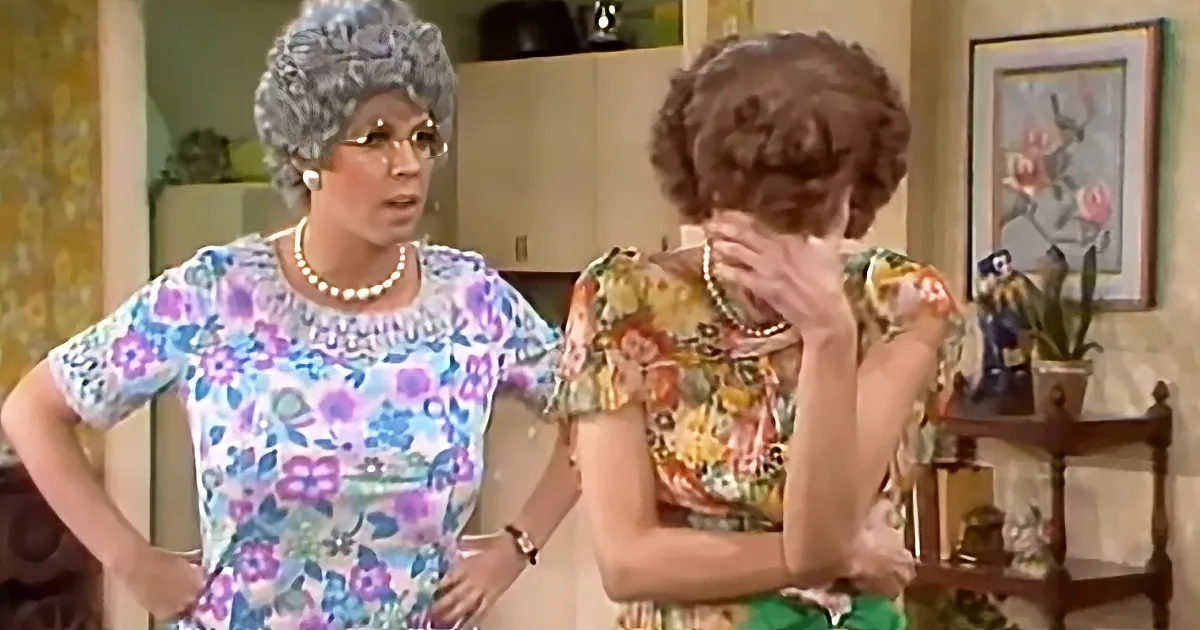 A woman in a floral dress with a pearl necklace stands with hands on hips, facing another woman in a similar dress who is...