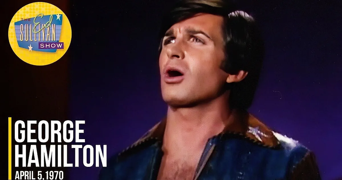 A man with a 1970s hairstyle and a brown jacket with blue accents is smiling.