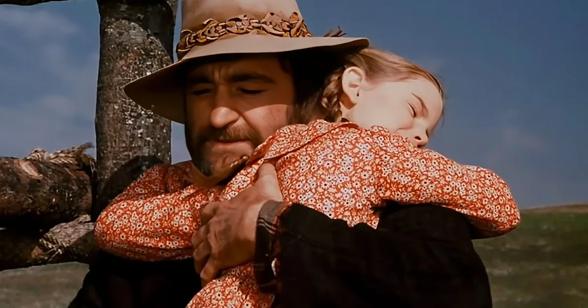 A man in a cowboy hat holds a sleeping child in a floral dress, set against a clear blue sky.