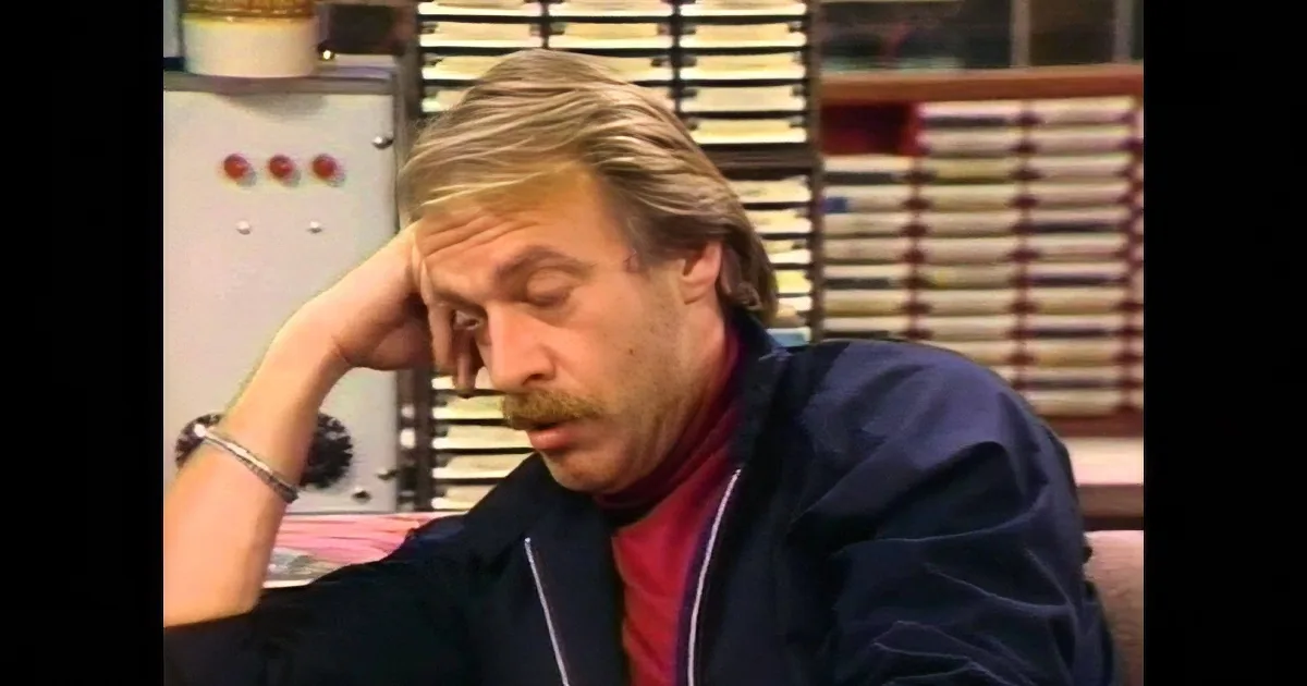 A man with a mustache speaks into a microphone in a studio setting.