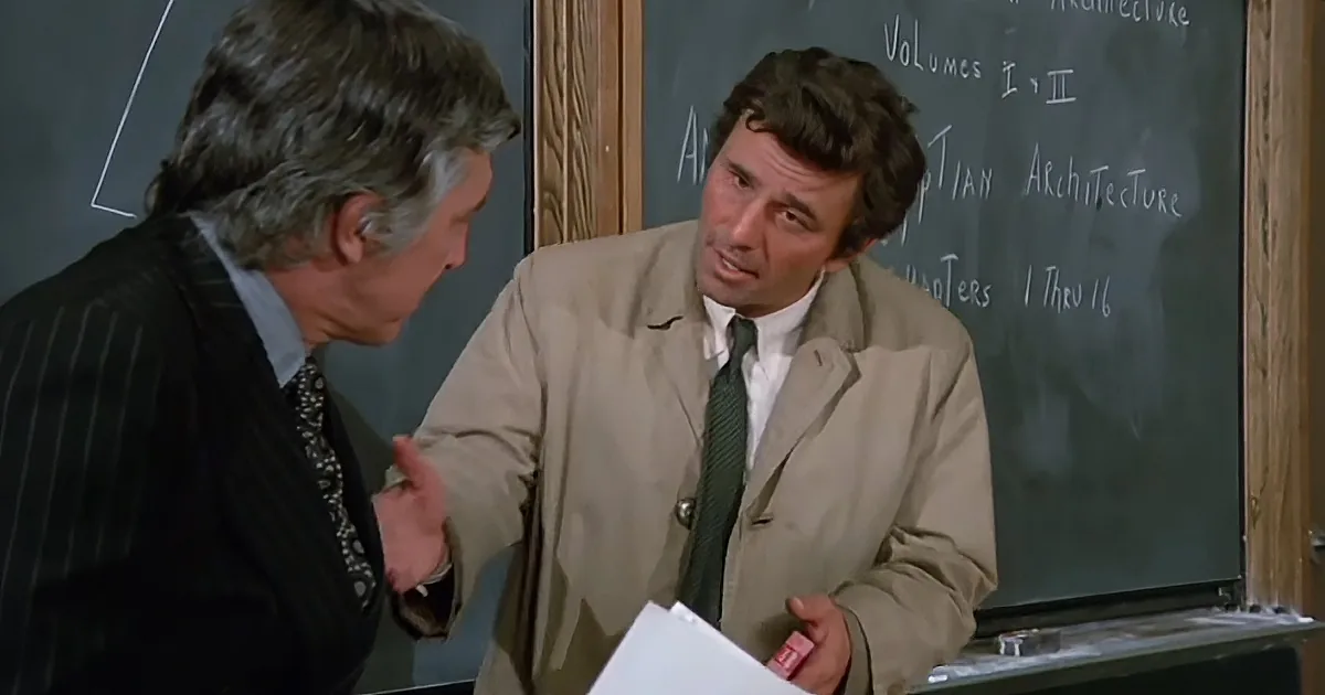 A man in a beige jacket holds papers, gesturing while speaking in front of a chalkboard.