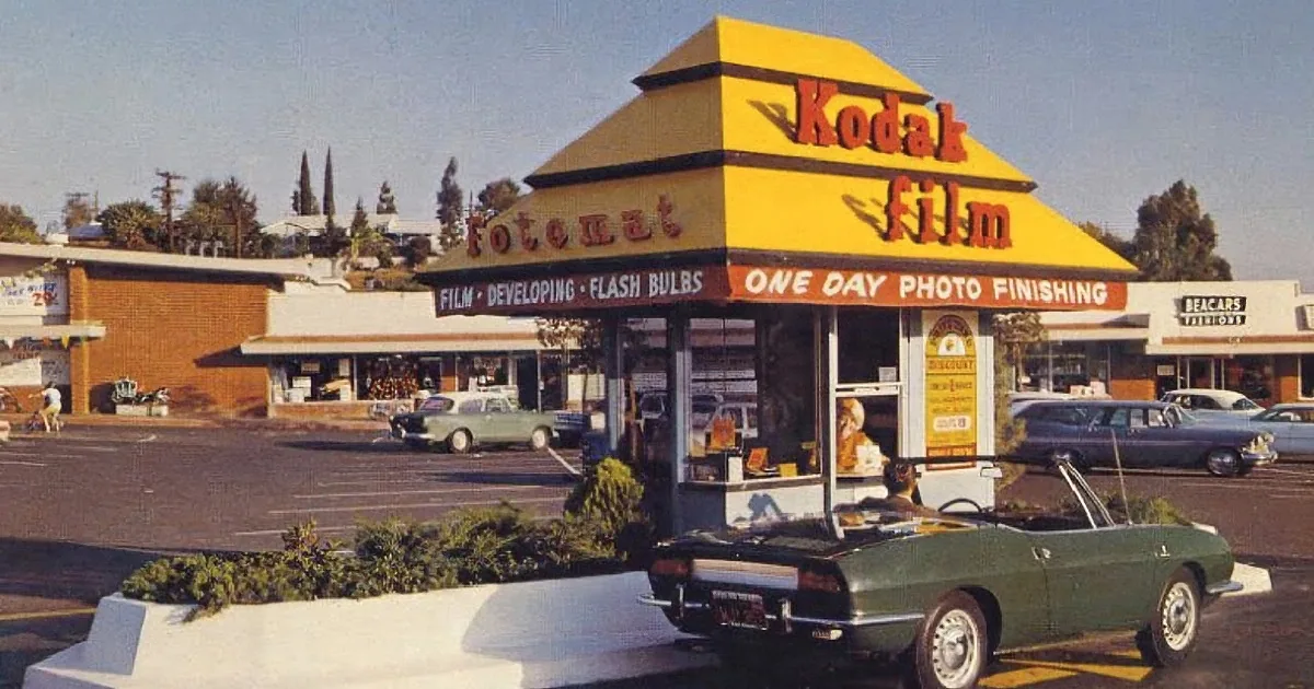 A vintage Kodak film store with a yellow and red sign, surrounded by classic cars and a parking lot.