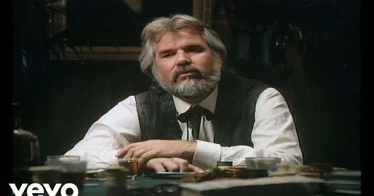 A man with a white beard and mustache, wearing a white shirt and dark vest, sits at a table.