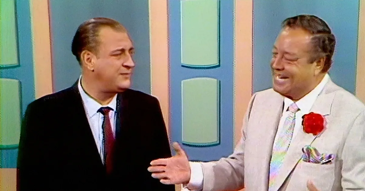 Two men in suits, one in black, the other in white, smiling and laughing in front of a colorful striped background.