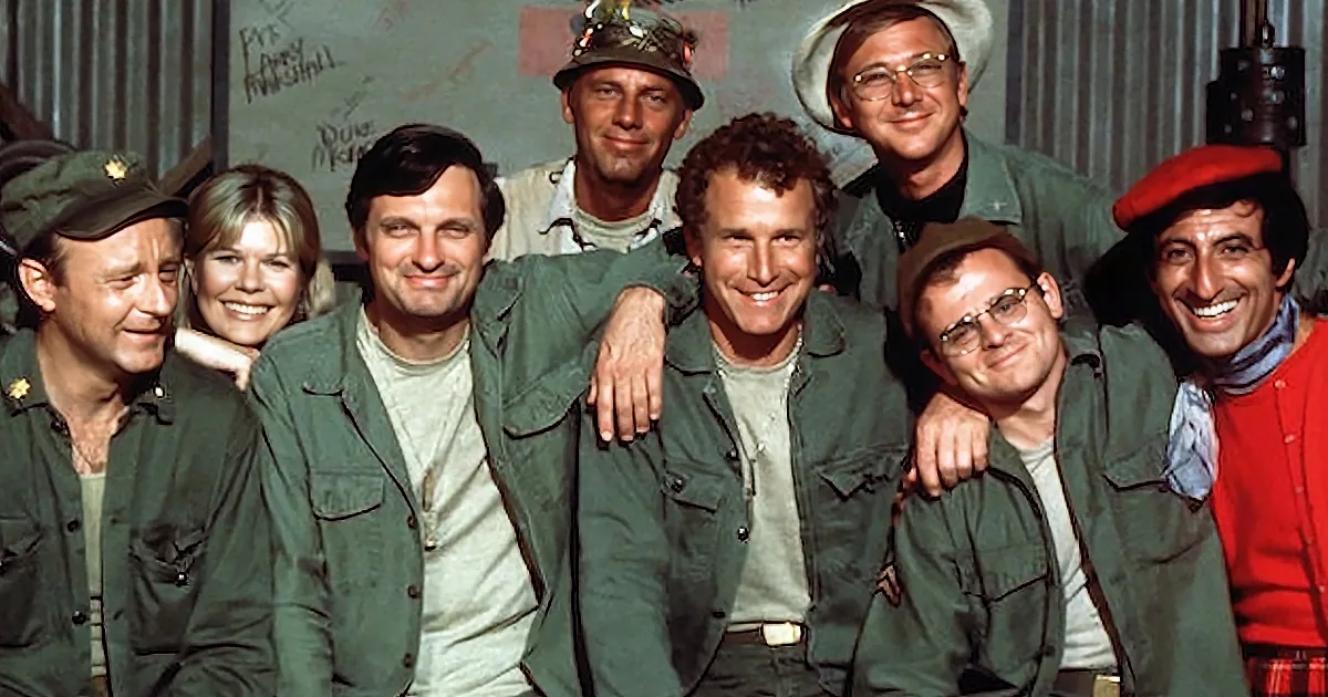 A group of actors in military-style uniforms smiles together, suggesting a camaraderie or team dynamic.