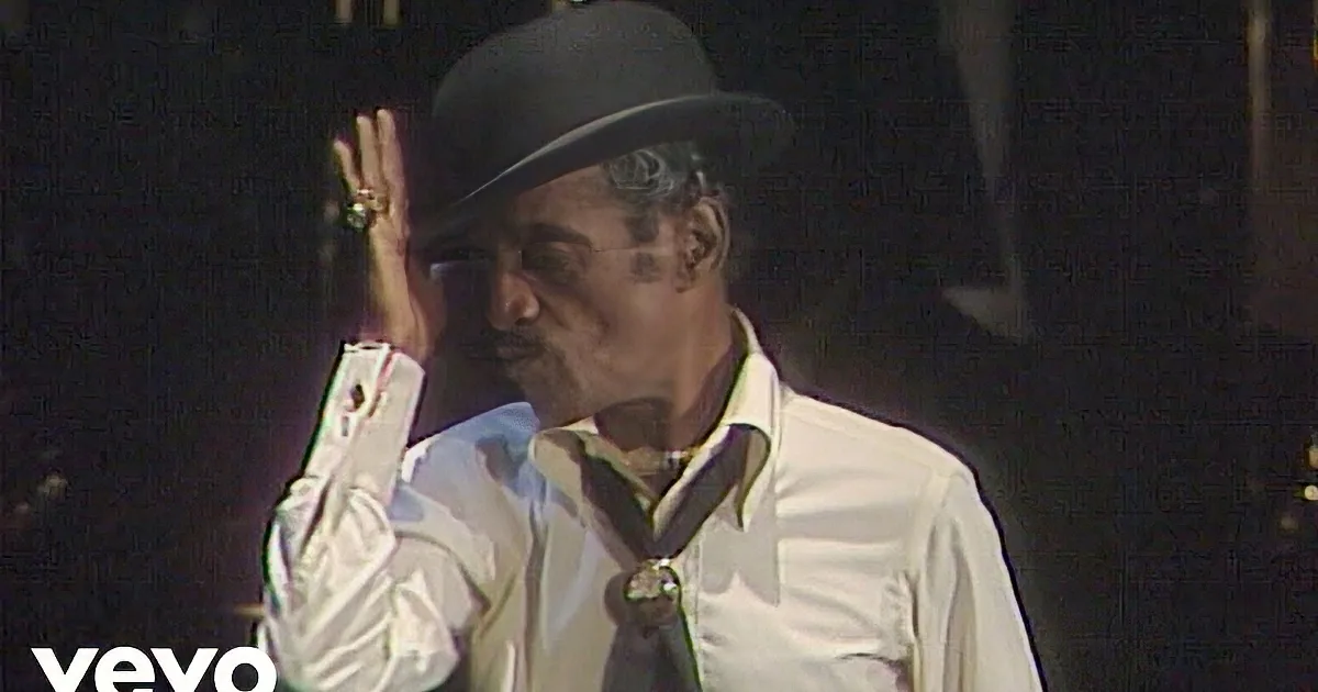A man in a white shirt and black hat holds a microphone, smiling, with a dark background and stage lights.