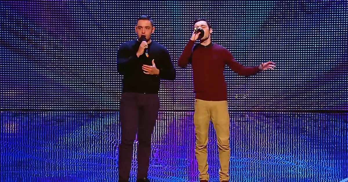 Two men on stage, one in black, one in red, holding microphones, performing in front of a LED screen.