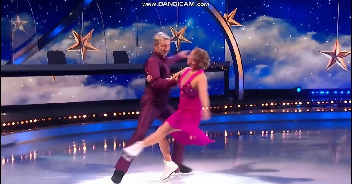 A couple in a dramatic skating pose, man in dark suit, woman in pink dress, on ice rink with blue stage lights.