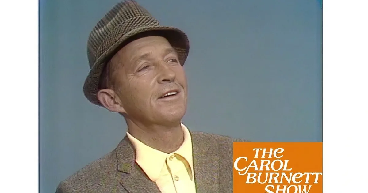 An older man wearing a brown hat, patterned suit, and yellow shirt with a black tie is speaking.