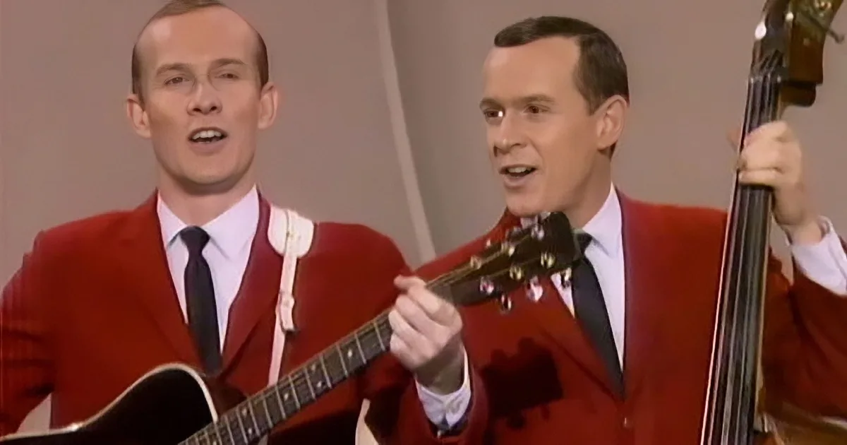 Two men in red suits play guitars and sing, one holding a bass.