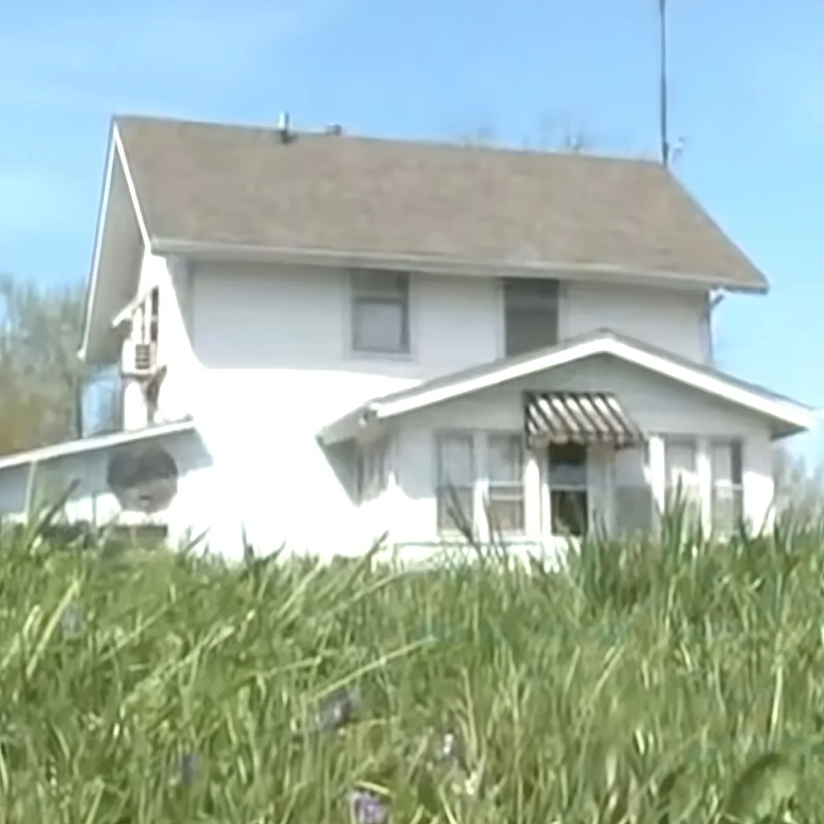 A white house with a brown roof sits amidst tall grass under a clear blue sky.