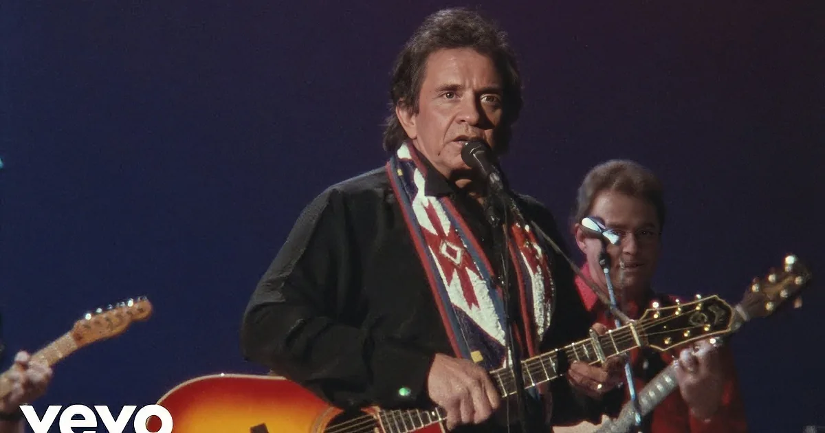 A man in a suit sings into a microphone while playing guitar, accompanied by other musicians on stage.