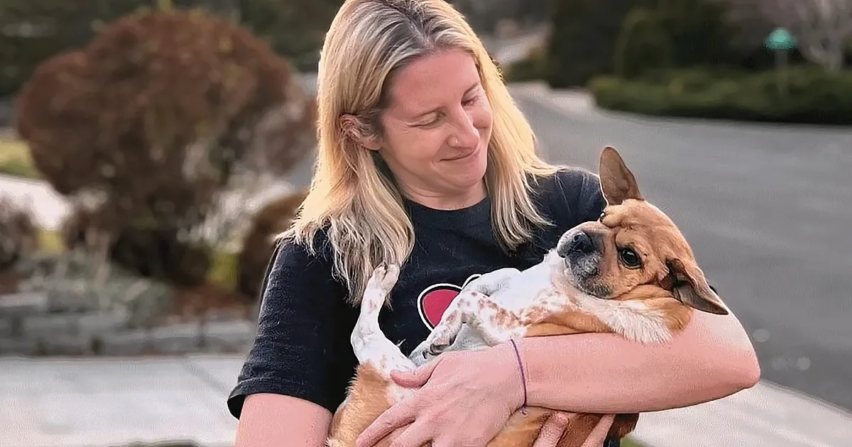 A woman with blonde hair holds a small brown and white dog, smiling outdoors.