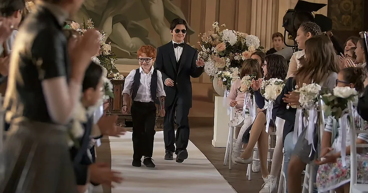 A man in a tuxedo walks down an aisle with a young boy, surrounded by flower girls and guests.