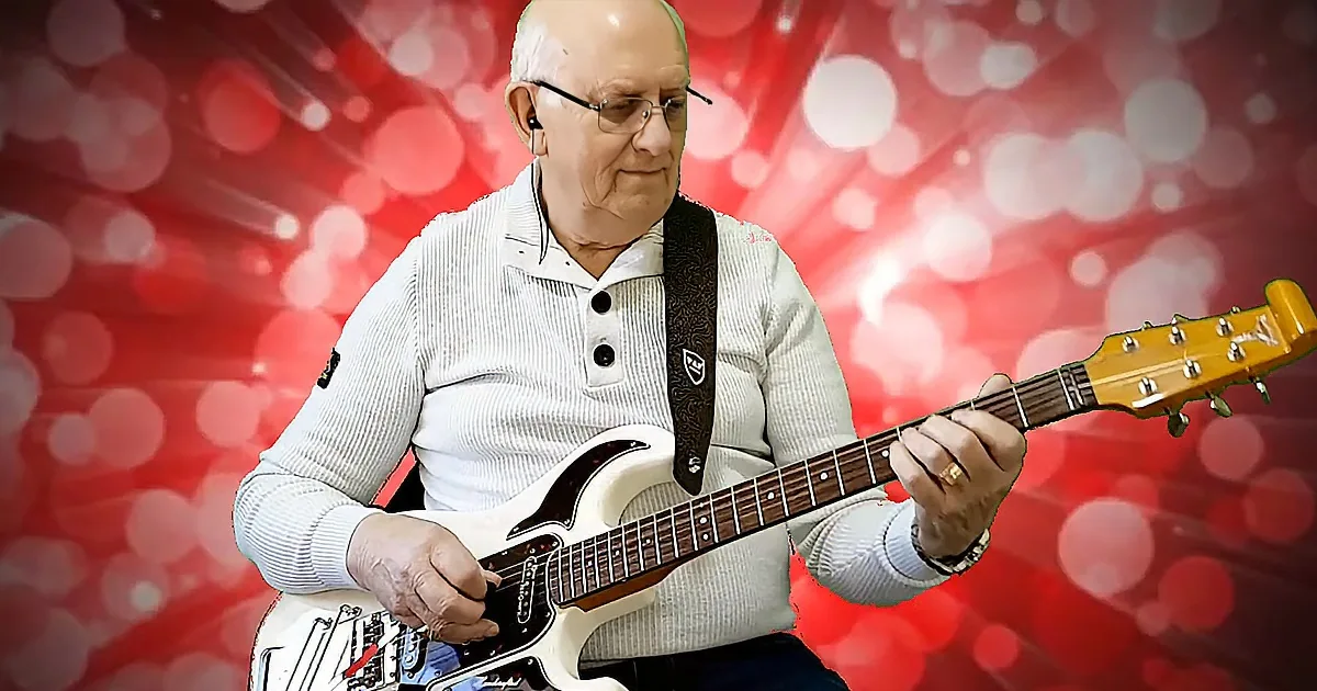 An elderly man plays a white electric guitar against a red, bokeh-lit background.