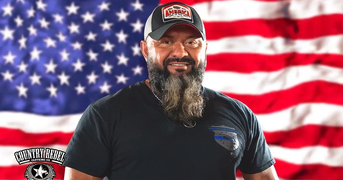 A man in a "Serve and Protect" shirt sings into a microphone, an American flag behind him.