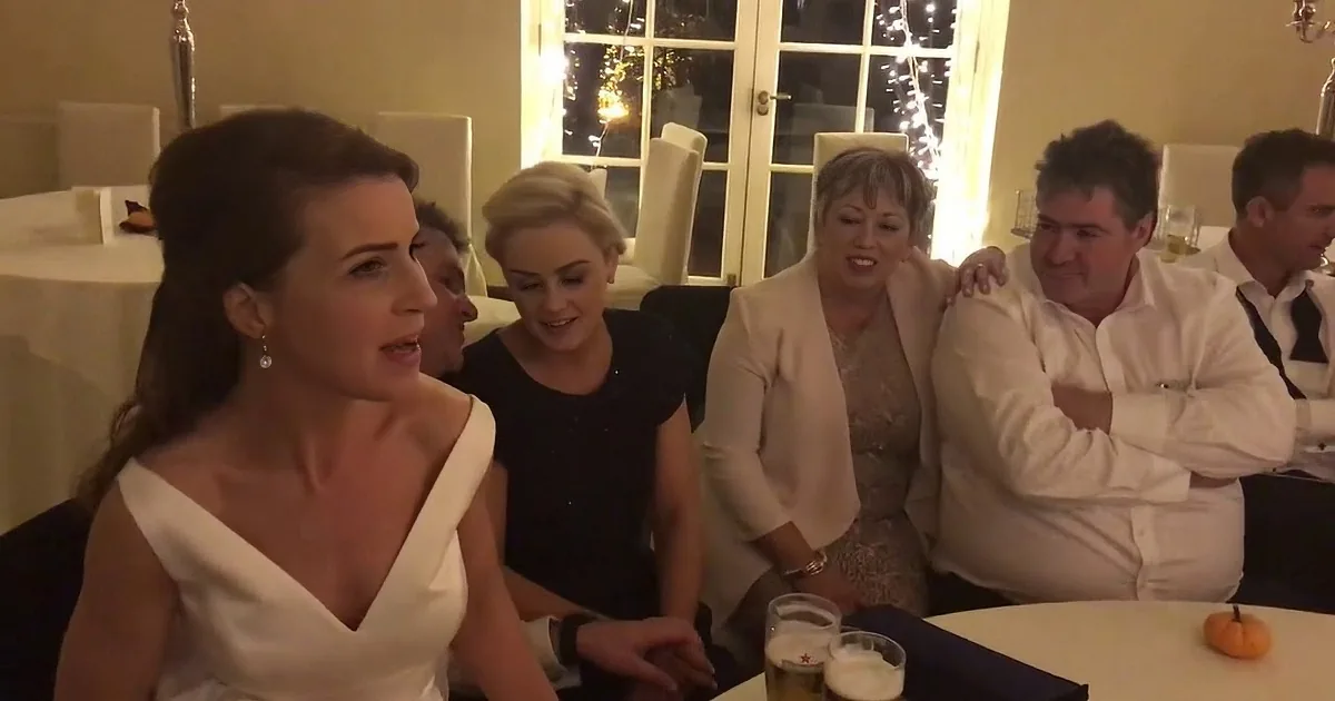 A bride in a white dress smiles at a group of seated guests at a formal event.
