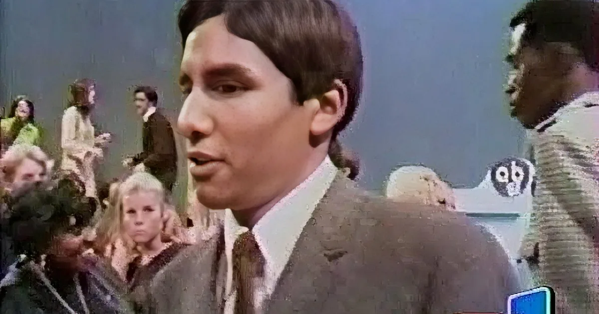A man in a brown suit is dancing on stage, surrounded by a group of people, with an audience watching and clapping.