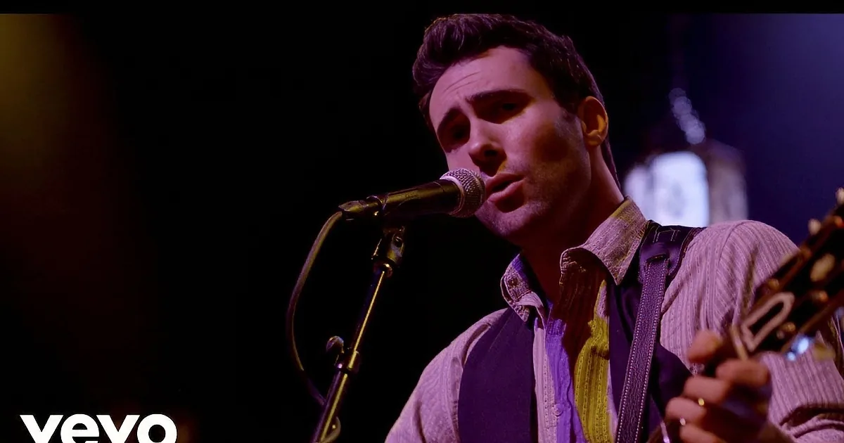 A man sings into a microphone while playing guitar on stage, surrounded by other musicians and an engaged audience.