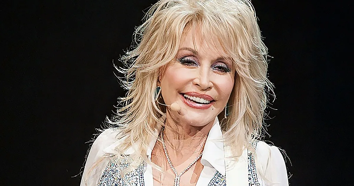 Dolly Parton smiles on stage, wearing a white shirt with a microphone and earrings.
