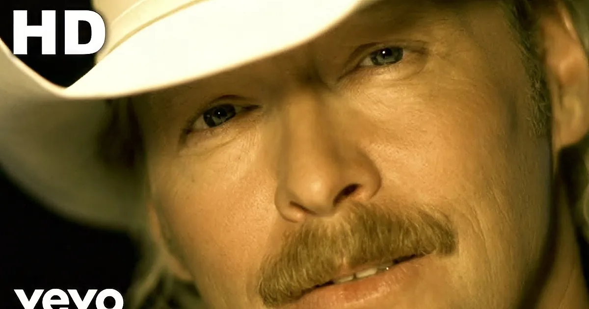 A man wearing a white cowboy hat and sunglasses, dressed in a tan suit, is seen against a teal background.