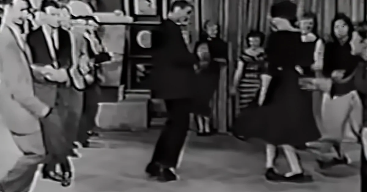 A man in a suit dances in a room with other people watching, dressed formally.