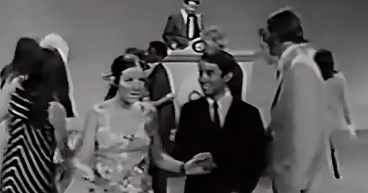 A man in a suit and tie is smiling and holding hands with a woman in a dress, surrounded by other people.