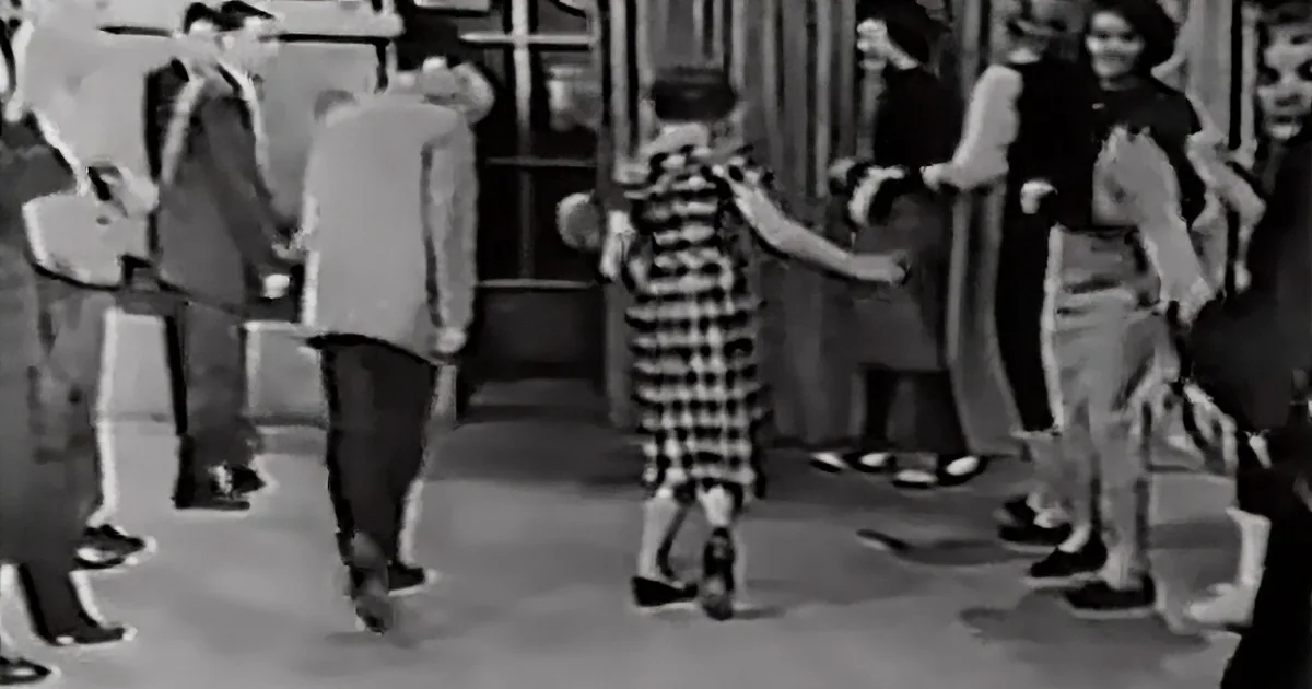 A young girl in a plaid dress dances in a crowd, surrounded by people in formal attire.