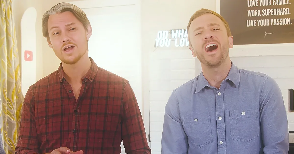 Two men, one in red plaid, the other in blue, appear to be singing or speaking in a room with motivational wall art.
