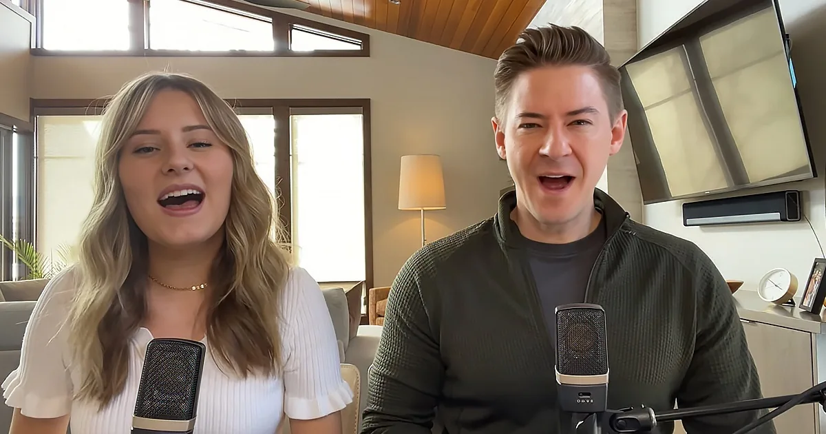 A man and woman are singing into microphones in a cozy room with a lamp and a clock in the background.
