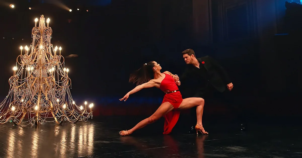 A couple in a dramatic dance pose, woman in red dress, man in black suit, illuminated by a grand chandelier.