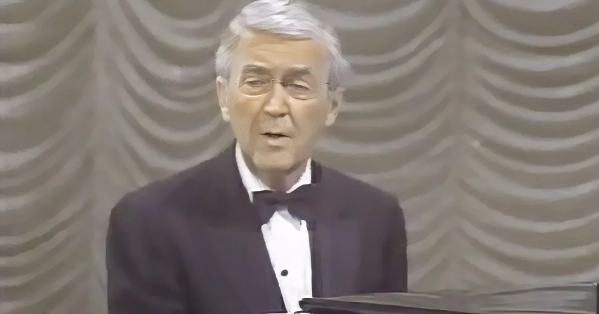 A man in a tuxedo stands at a podium, speaking in front of a patterned backdrop.