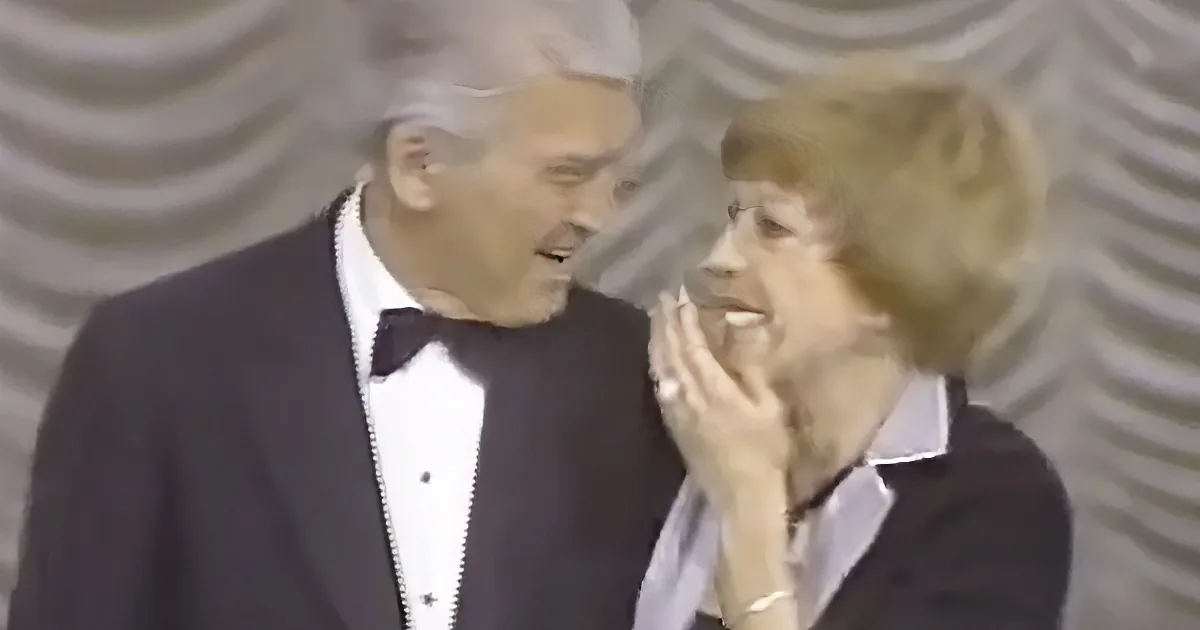 A man in a tuxedo whispers into a woman's ear, both appearing engaged in a private conversation.