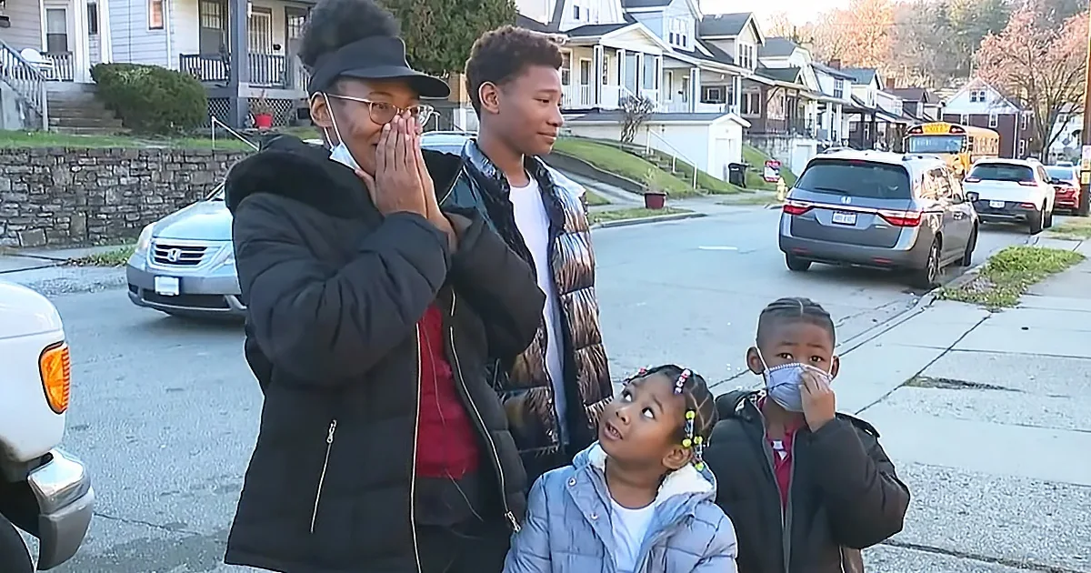 A woman in a black jacket and hat stands with children on a suburban street, covering her face with her hands.