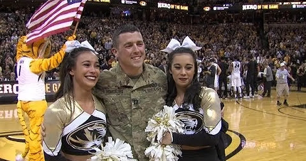A soldier in camouflage hugs two cheerleaders in a gymnasium, surrounded by cheering spectators.