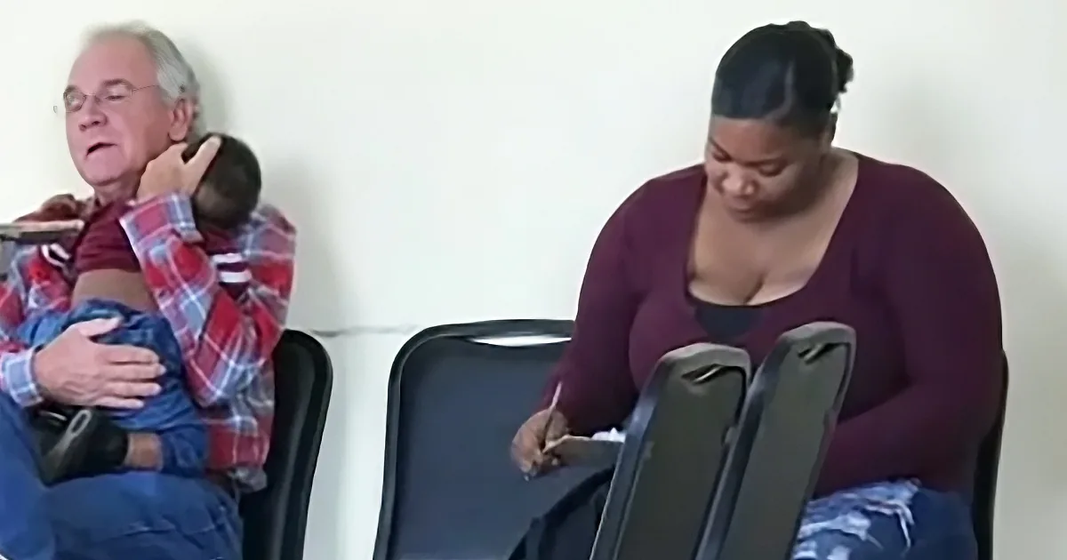 A woman in a maroon top is sitting and writing on a clipboard, while a man in a plaid shirt holds a baby.