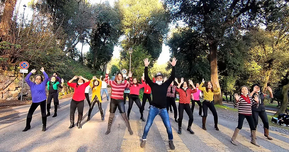A group of people in colorful clothing are dancing on a road with trees and a stone wall in the background.