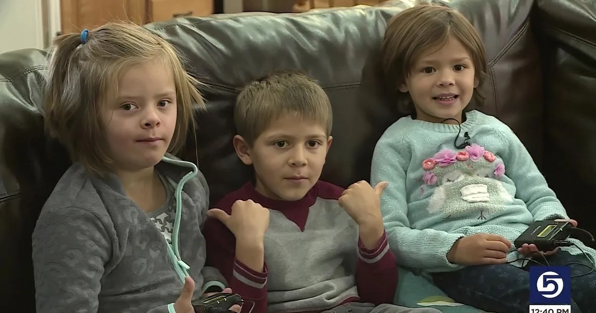 Three children sit on a couch, one giving a thumbs-up, all looking at the camera.