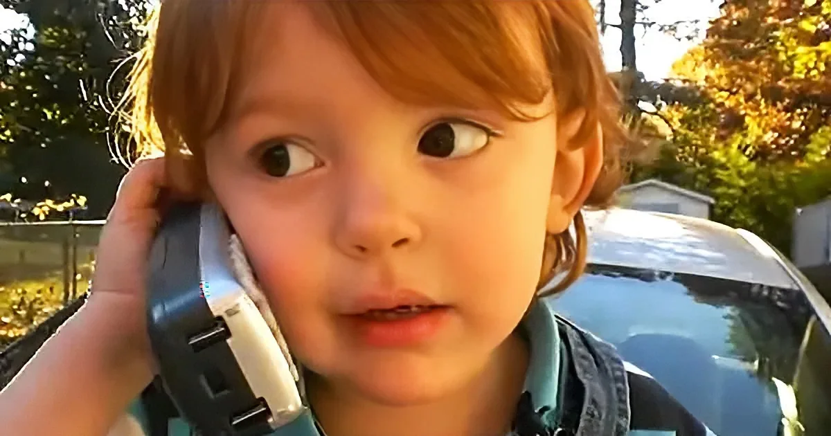 A young child with red hair holds a toy phone to their ear, looking to the side with a curious expression.