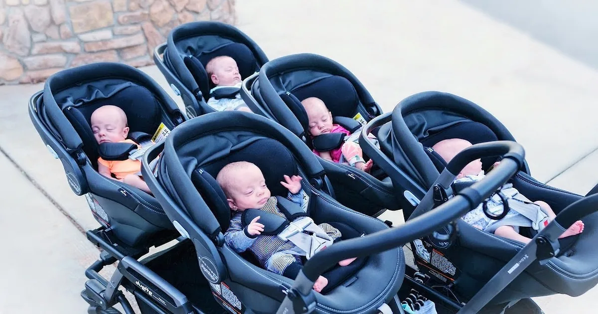 Five babies in strollers lined up on a sidewalk, all facing forward.