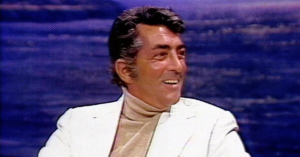 Two men are seated at a table, one in a white suit, the other in a plaid shirt, engaged in conversation.