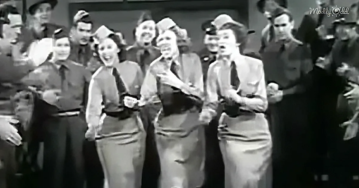 Three women in uniform sing with a group of men in military attire.